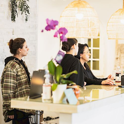 Three people are at a reception desk, with two women behind it and one in front of it. There are hanging lights and a potted orchid.