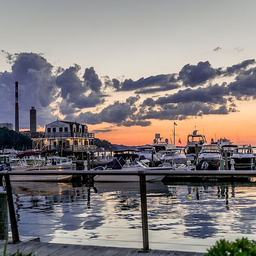 The image shows a marina at sunset with boats docked and industrial chimneys in the background, reflecting on the calm water, all under a cloudy sky.