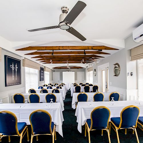 The image shows a conference room setup with rows of blue and gold chairs and white covered tables. A projector screen is at the front.