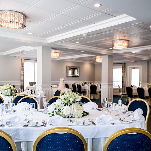 The image shows a decorated banquet hall with round tables covered in white cloth, blue chairs, floral centerpieces, and elegant lighting fixtures.