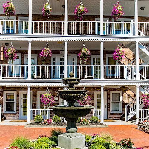 The image shows a three-story building with white railings and numerous hanging flower baskets. A decorative fountain is in the foreground, surrounded by greenery.