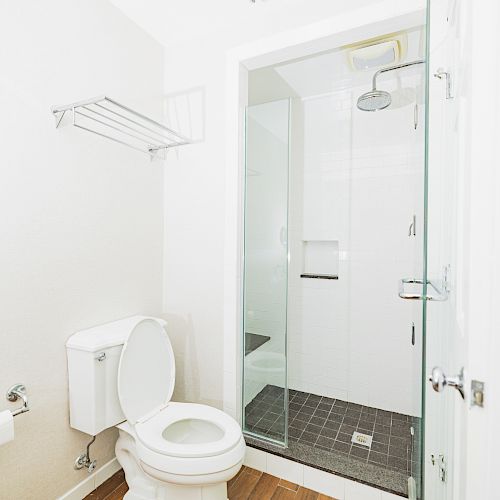 The image shows a bathroom with a toilet, a shower with a glass door, a toilet paper holder, and an empty shelf on a white wall.