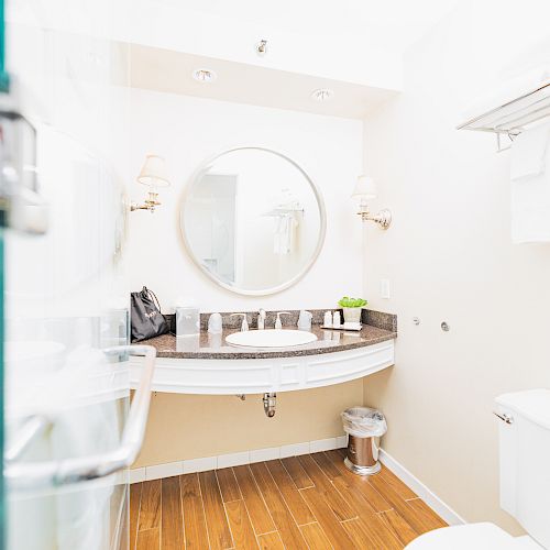 A bright bathroom with a round mirror, sink, and towels hanging on a rack. The floor has wooden panels and there’s a trash can under the sink.