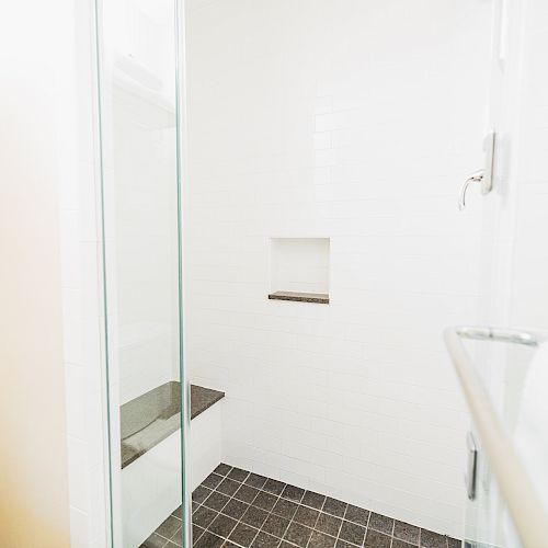 This image shows a white-tiled shower area with a glass door and a bench. There’s a built-in shelf on the wall and dark floor tiles.