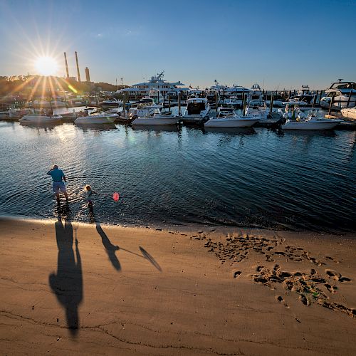 A person and a child are standing on a sandy beach near the water, with boats docked in the marina and a setting sun in the background.