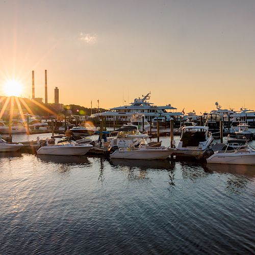 The image shows a marina at sunrise or sunset, with numerous boats docked in the calm water and industrial structures in the background.