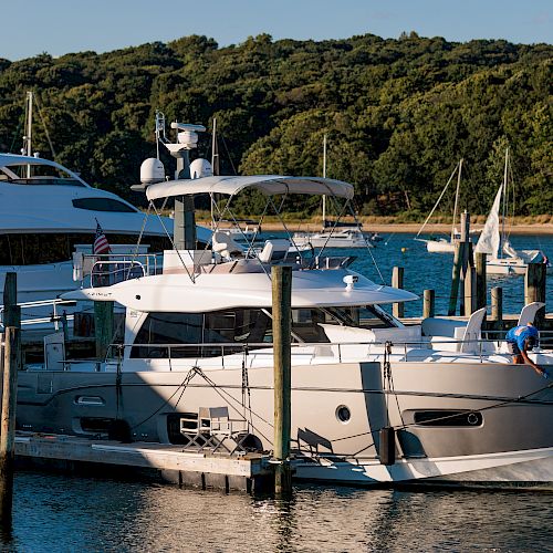 A marina with several luxury yachts docked, surrounded by water and greenery; a person cleans one of the yachts.