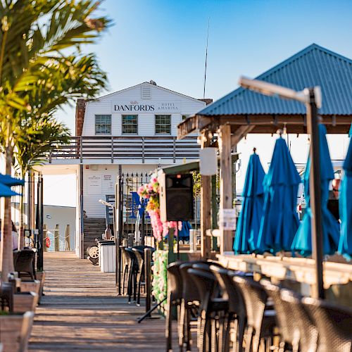 An outdoor seating area with tables, chairs, and blue umbrellas, possibly a restaurant, leading to a building, with palm trees in the background.