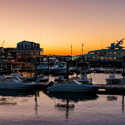 A tranquil marina at sunset with boats docked, a large yacht, and industrial buildings in the background, reflecting on calm waters.