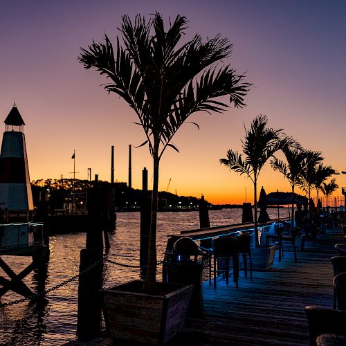 A sunset view of a waterfront with palm trees, a lighthouse, and an outdoor seating area along a wooden dock, illuminated with soft lighting.