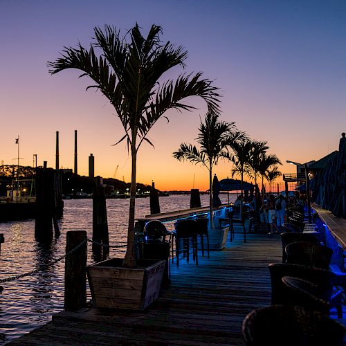 A waterfront scene at sunset with palm trees, a lighthouse, seating area, and string lights creating a relaxing ambiance along the pier.