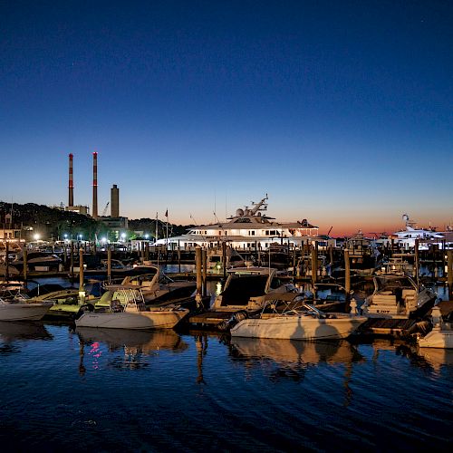 A marina at dusk with boats docked in calm waters, buildings, and smokestacks in the background under a twilight sky, fully lit up.