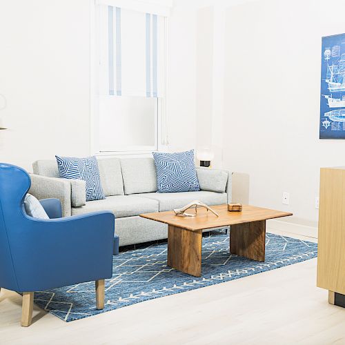 A modern living room with a gray sofa, blue armchair, wooden table, and a nautical-themed wall art. The room has a bright, minimalist style.