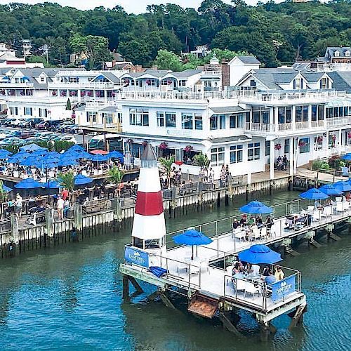 This image shows a waterfront scene with a white and red lighthouse, outdoor dining areas with blue umbrellas, and buildings by the water's edge.