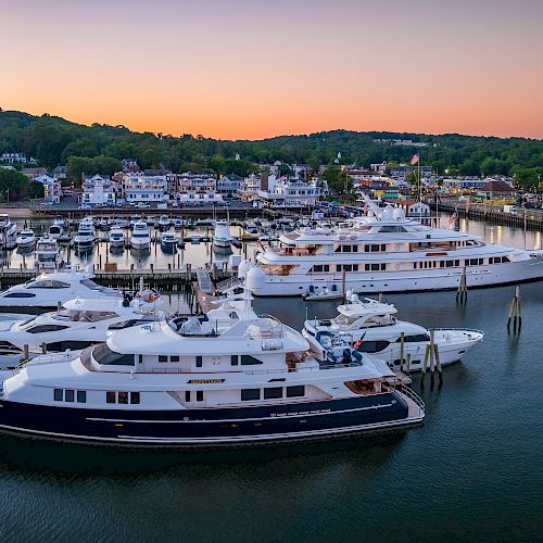 The image shows a marina at sunset with several luxury yachts docked. The background has a scenic view of hills and a small town.