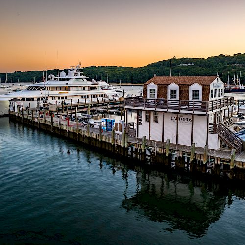 A marina at sunset with a large yacht docked alongside a pier and a building overlooking the water and boats.