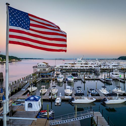 A marina during sunset with numerous boats docked, an American flag prominently displayed in the foreground, and a calm, picturesque waterfront.