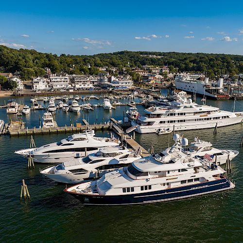 The image shows a marina with several large yachts docked near a coastal town, set against a backdrop of trees and buildings in clear weather.