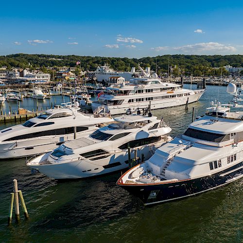 Several luxury yachts are docked in a marina with a small coastal town in the background and a clear blue sky above.