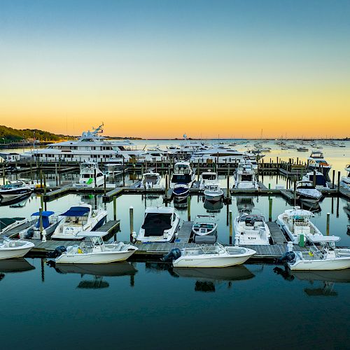 A marina filled with various boats, with an industrial facility and hills in the background during a picturesque sunset over the calm water.