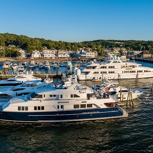 Multiple yachts are docked at a marina with a coastal town and greenery in the background, under a clear blue sky.