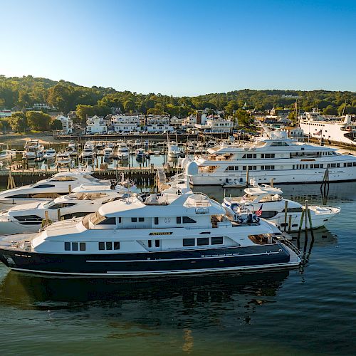 Several yachts are docked at a marina, surrounded by calm waters and a nearby town with lush, green hills in the background, under a clear blue sky.