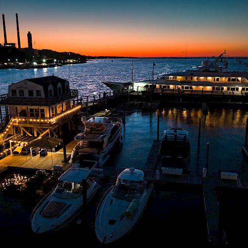 A marina at sunset with boats docked, a lit-up building, and industrial chimneys silhouetted against the vibrant evening sky.