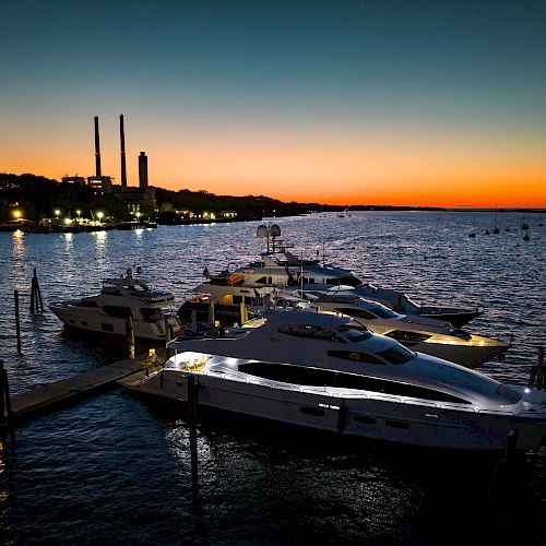 A marina at sunset with several yachts docked, industrial structures visible in the background, and a serene orange and blue sky.