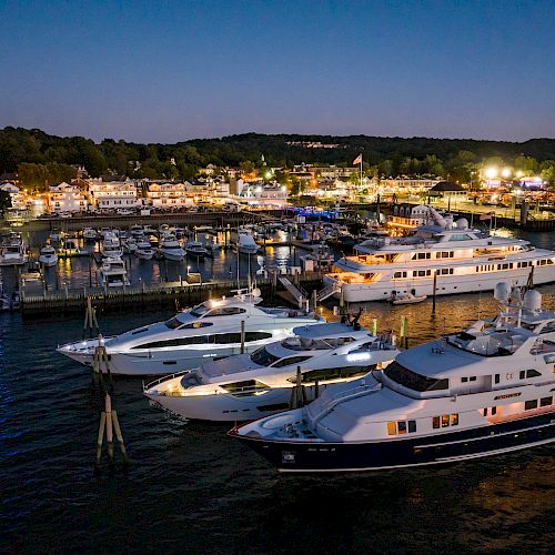 A marina at dusk with several yachts docked, buildings illuminated in the background, and a wooded area silhouetted against the twilight sky.