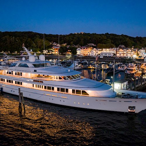 A large, well-lit yacht docked at a marina during the evening, with buildings and trees visible in the background, under a darkening sky.