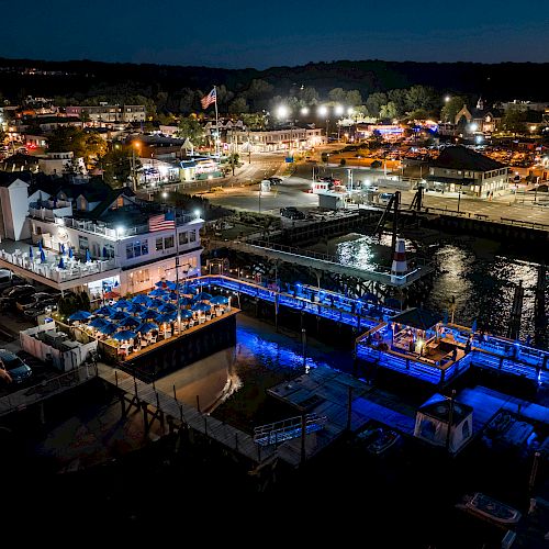 This image depicts a nighttime view of a lively waterfront area with illuminated structures, restaurants, and boats docked along a pier.