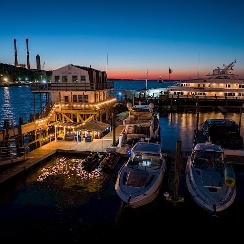 A serene harbor scene at dusk, featuring boats docked near a lit-up building, with a beautiful sunset in the background and calm water.