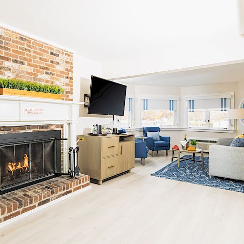 A cozy living room features a brick fireplace, wall-mounted TV, modern furniture, and large windows offering natural light.