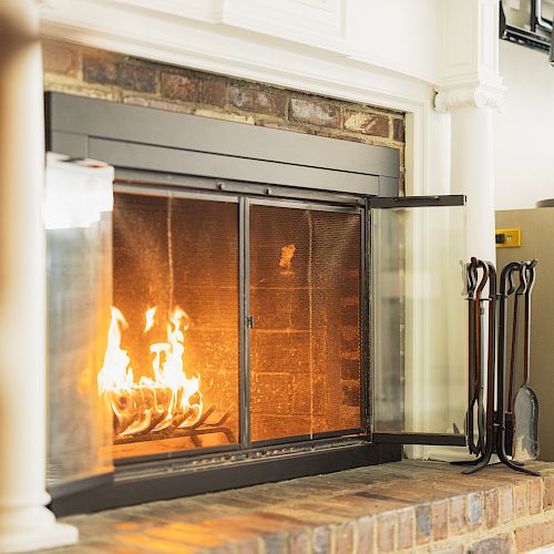 A cozy, modern living room features a glass-enclosed fireplace with a fire burning, a toolset nearby, and a cabinet with candles on top.