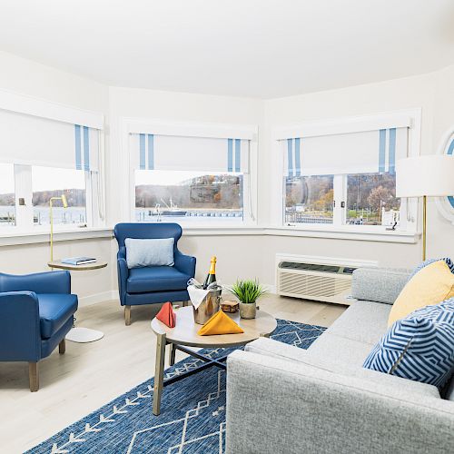 A cozy living room with blue chairs, a gray sofa, a coffee table with drinks and snacks, a blue rug, and large windows with striped blinds.