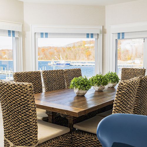 A dining area with a wooden table, wicker chairs, and potted plants. Large windows offer a scenic view of a waterfront and autumn foliage outside.