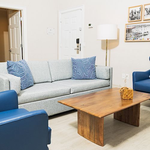 A cozy living room featuring a gray sofa with blue pillows, blue armchairs, a wooden coffee table, and wall art. A door and lamp are visible.