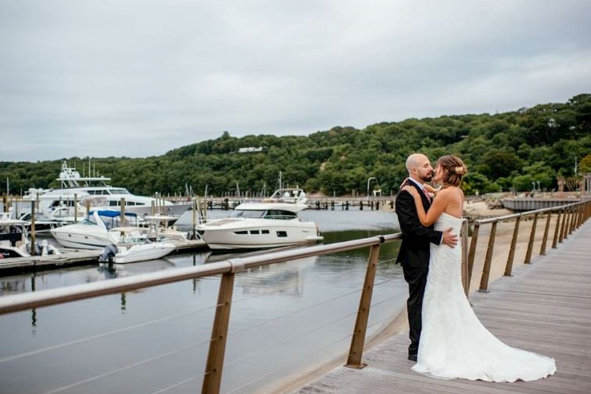 A bride and groom embrace on a dock with several yachts moored in the background and lush green hills behind.