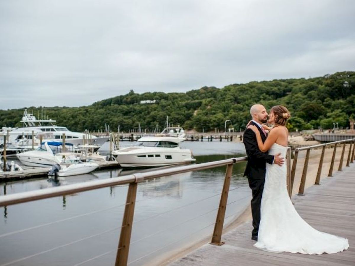 A bride and groom embrace on a dock with several yachts moored in the background and lush green hills behind.