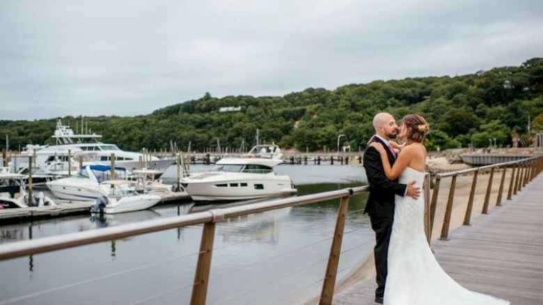 A bride and groom embrace on a dock with several yachts moored in the background and lush green hills behind.