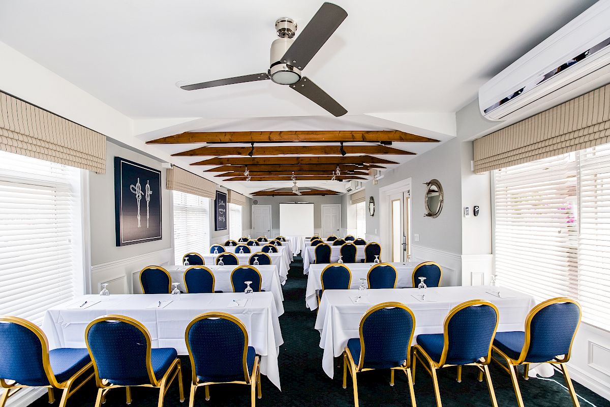 A conference room with blue chairs, white tablecloths, fans, air conditioning, and a screen at the front, set up for a meeting or presentation.
