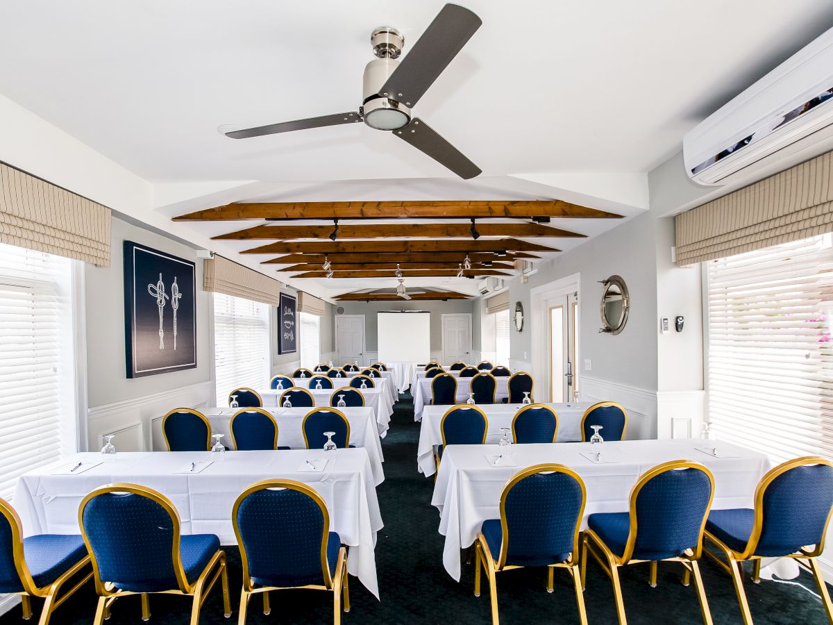 A conference room with blue chairs, white tablecloths, fans, air conditioning, and a screen at the front, set up for a meeting or presentation.