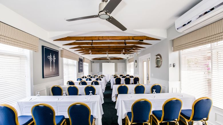 A conference room with blue chairs, white tablecloths, fans, air conditioning, and a screen at the front, set up for a meeting or presentation.