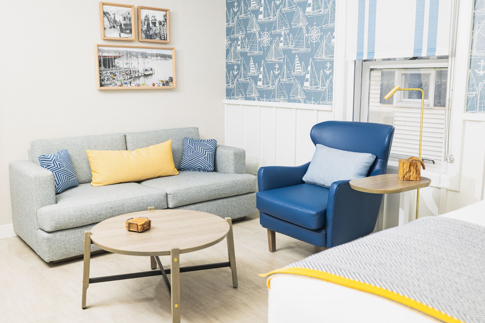 A cozy and stylish living room with a gray sofa, blue armchair, round coffee table, framed pictures on the wall, and nautical-themed wallpaper.