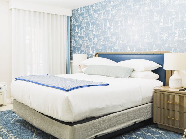 The image shows a neatly made bed in a well-decorated bedroom with nautical-themed wallpaper, bedside tables, and soft ambient lighting.