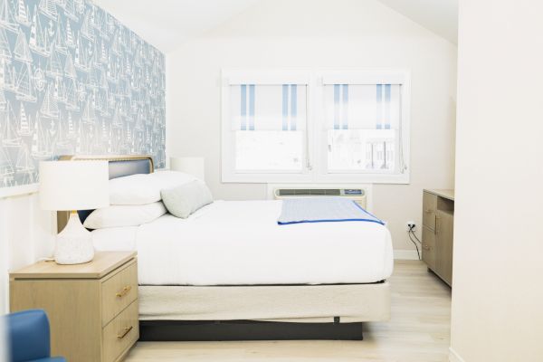 A clean, bright bedroom with a bed, bedside tables with lamps, a dresser, and windows with striped blinds. The walls have a nautical-themed wallpaper.