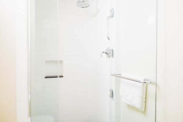 The image shows a clean, white tiled shower with a glass door, a towel on the handle, a showerhead, and a built-in shower shelf.