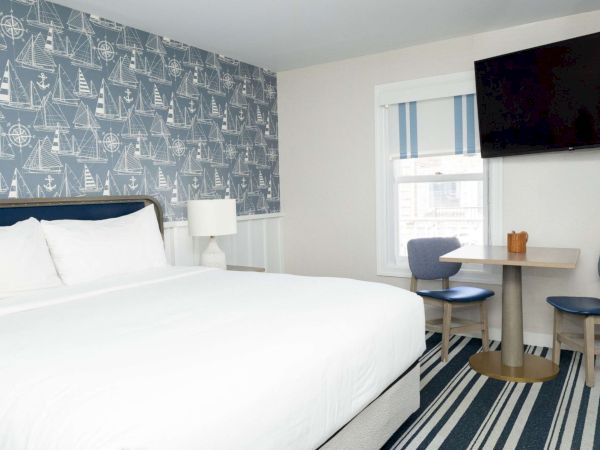 A bright hotel room with a large white bed, nautical sailboat wallpaper, a desk by a window, and a wall-mounted TV.