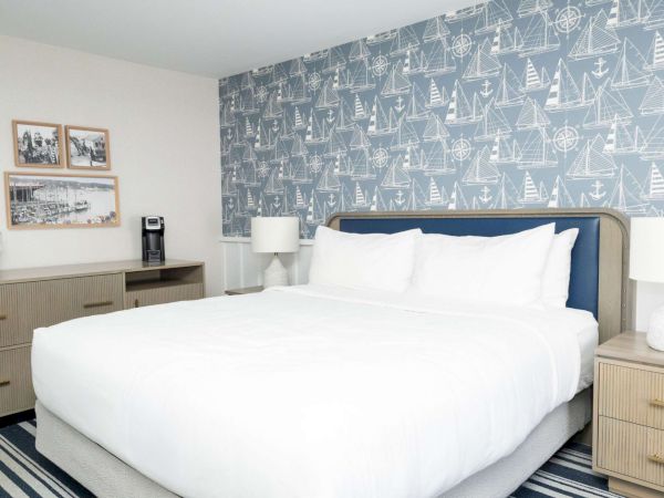 A bright hotel bedroom with a large white bed, nautical sailboat wallpaper, and framed photos on the wall.