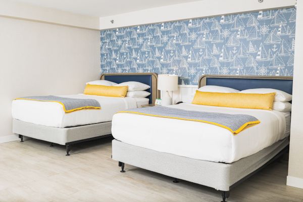 The image shows a modern hotel room with two double beds, blue and white nautical-themed wallpaper, and yellow accents on the bedding, ending the sentence.